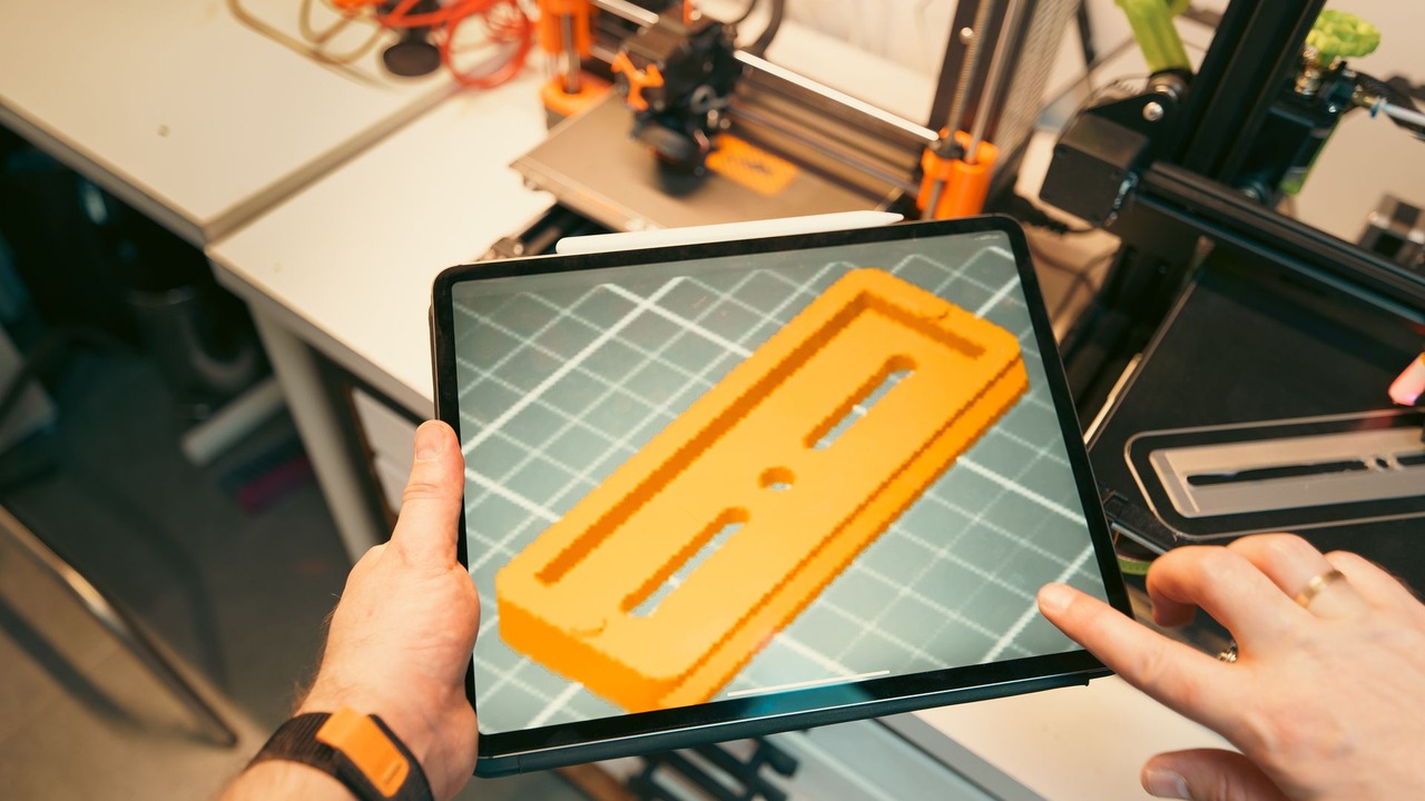 Una persona consulta un modelo 3D en una tableta frente a impresoras y prototipos en un taller.