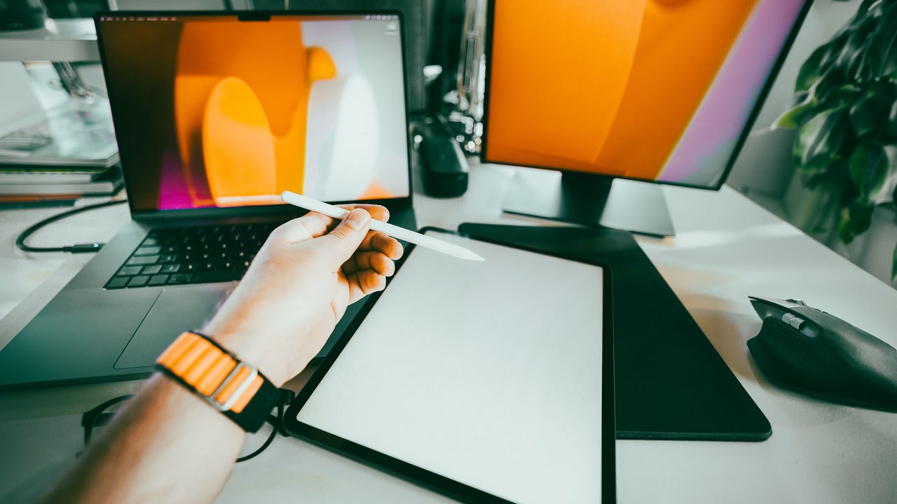 Mano con stylus sobre una tableta gráfica junto a un portátil y un monitor.