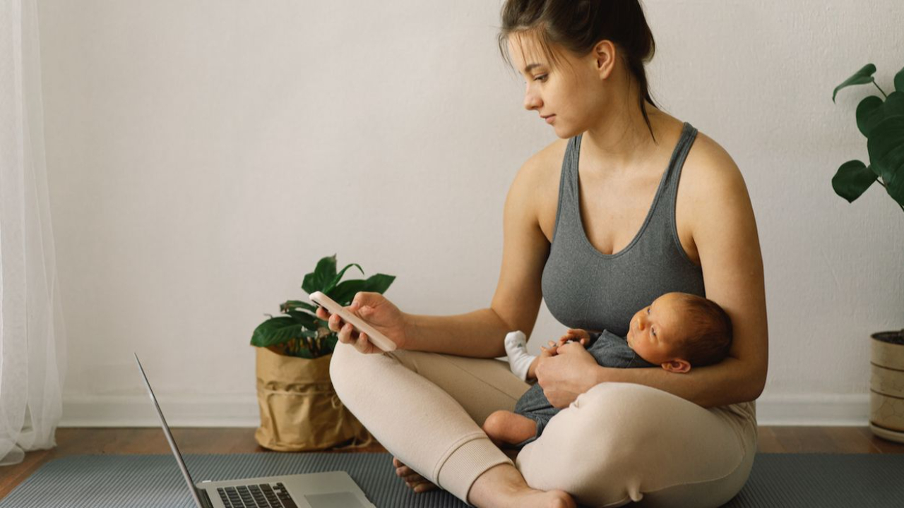 young woman sitting on yoga mat with an open laptop, holding an iphone in one hand and baby in the other