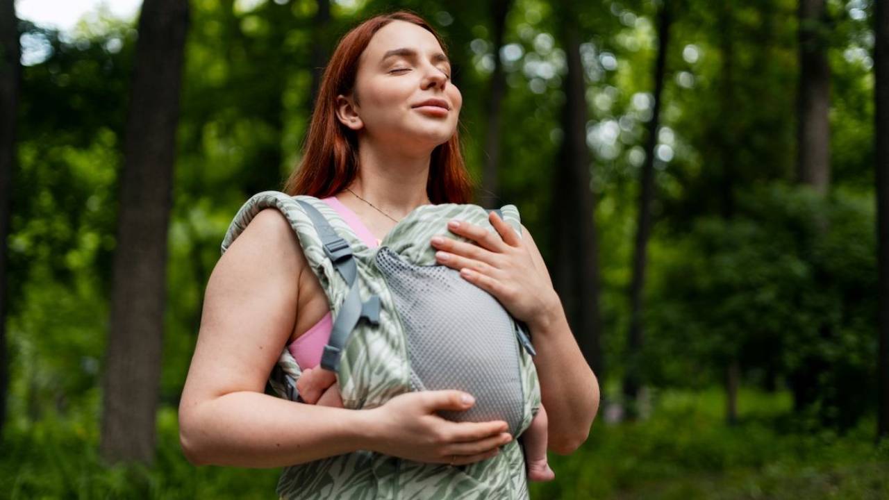 mother holding newborn in front carrier surrounded by green trees
