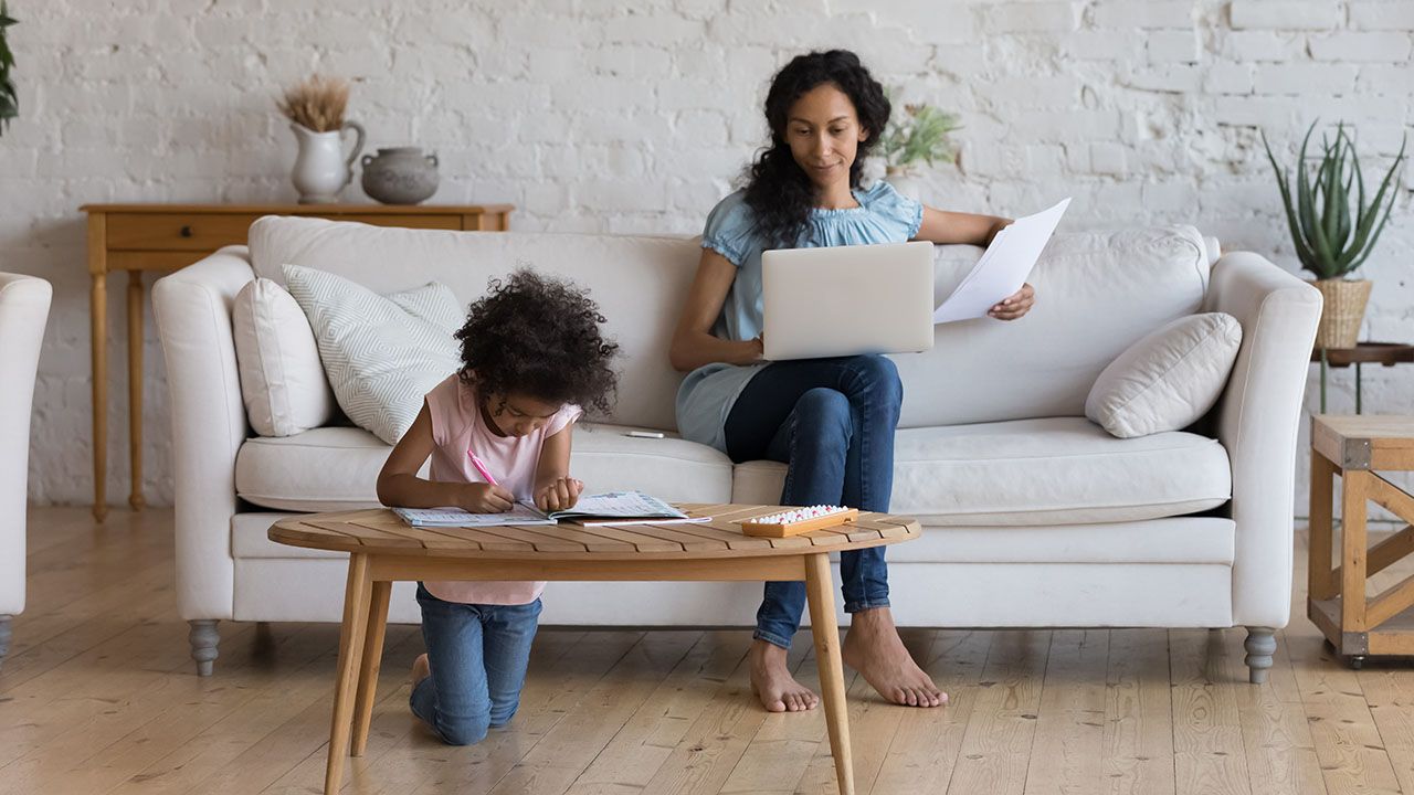 Adult using a laptop on a sofa while a child works at a nearby table