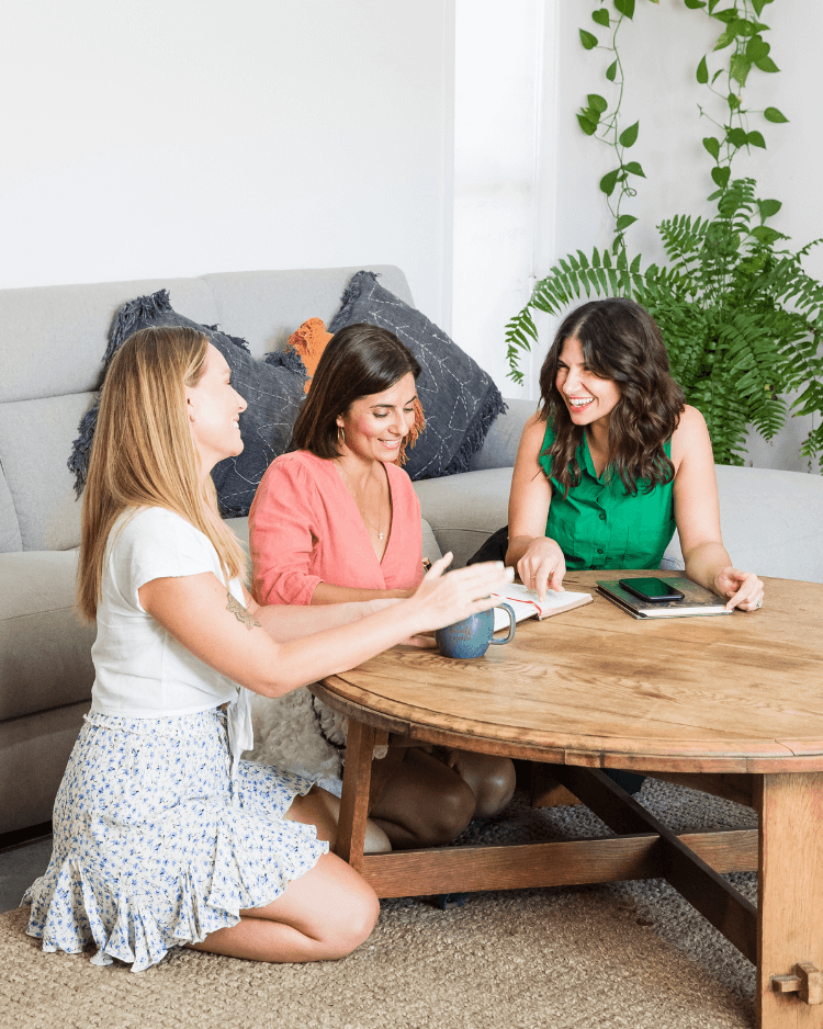 Three women kneeling at a coffee table smiling and laughing. Working together and building business marketing.