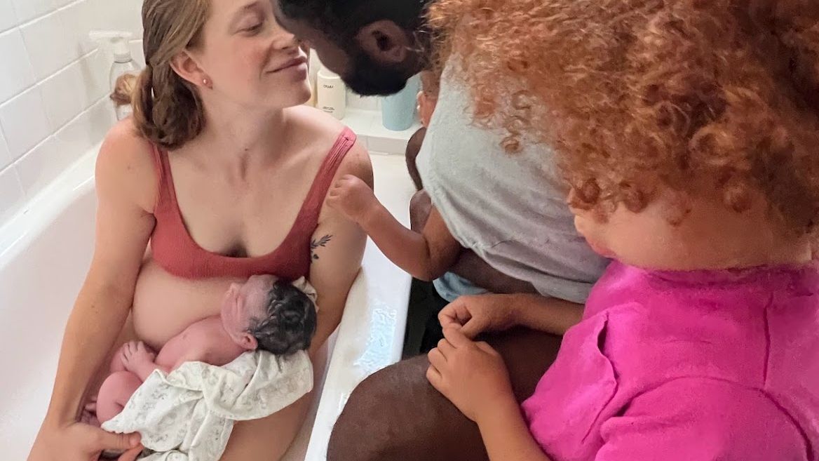 woman holding baby in bathtub after giving birth, leaning over to kiss man