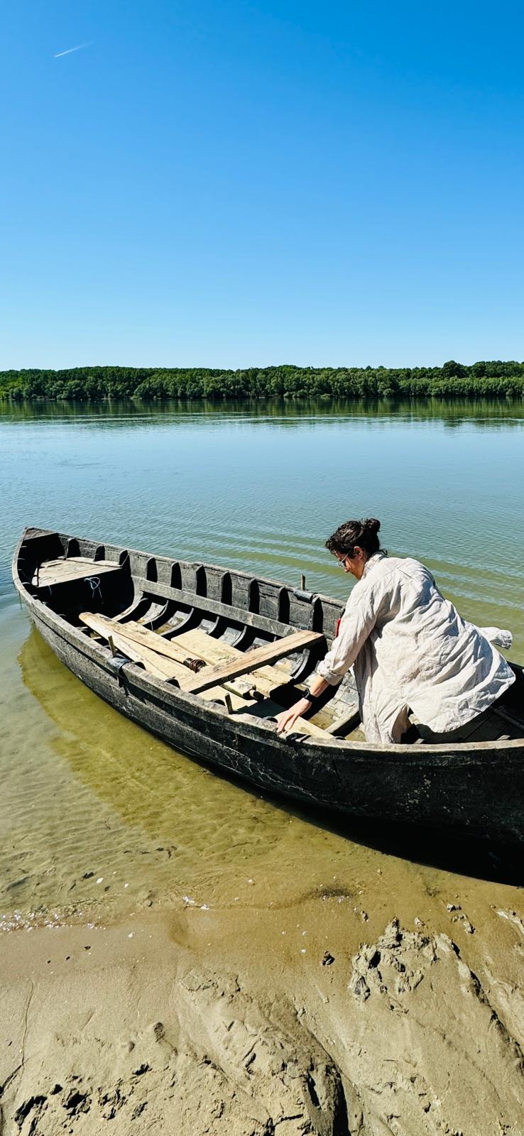 Elena Visentin china su una vecchia barca di legno in riva al fiume, cielo azzurro e acqua calma sullo sfondo