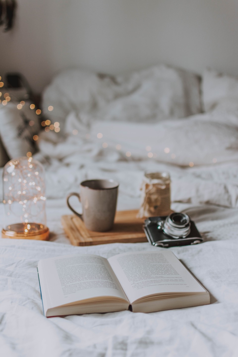 A cozy bed with an open book, a warm mug and soft holiday lights