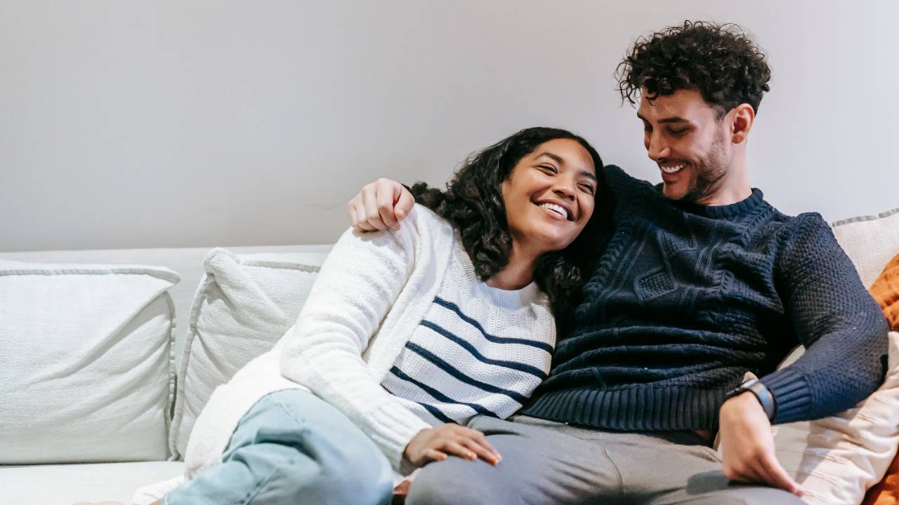 Couple sitting on a couch, smiling and laughing while looking at each other, with the man's arm around the woman.