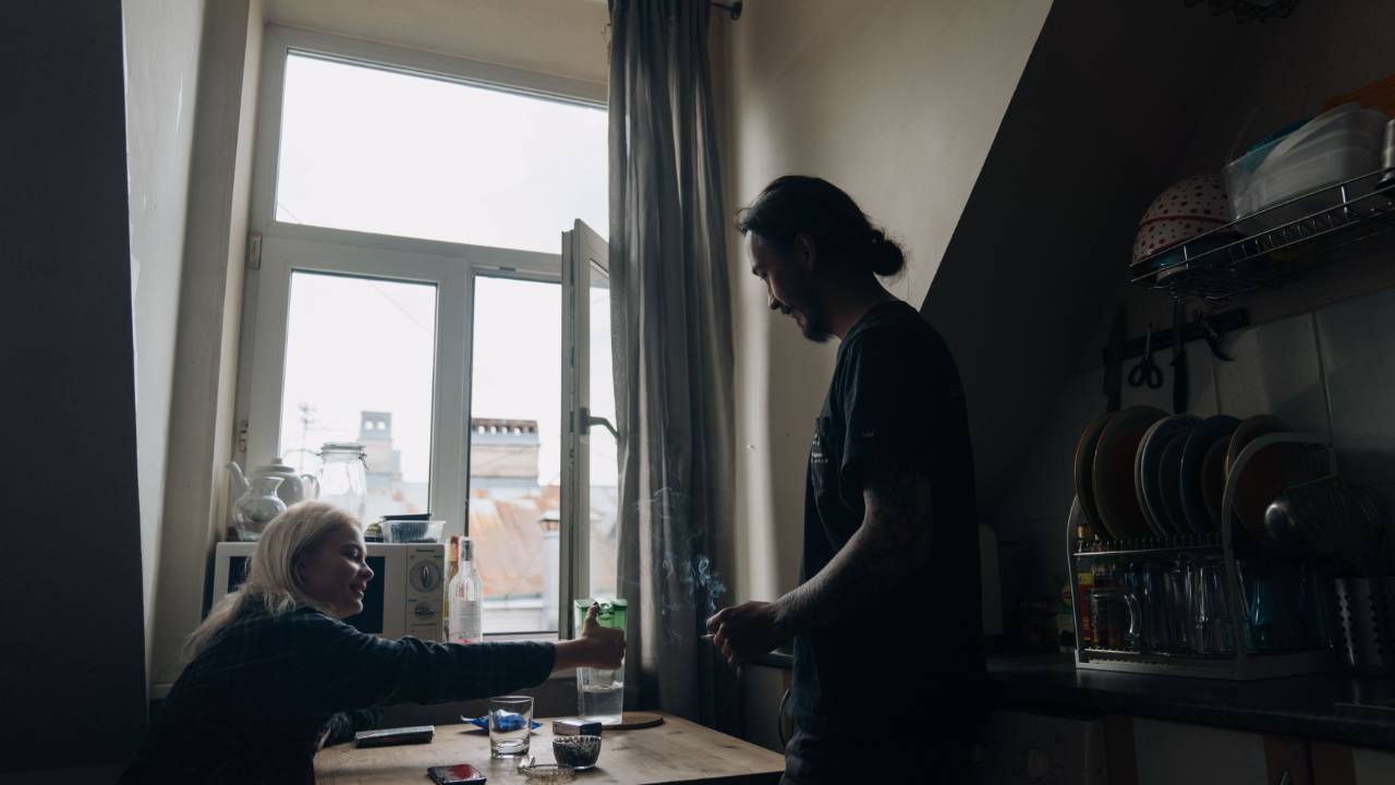 Happy couple in their apartment kitchen, with the man standing and the woman seated and reaching for a water pitcher, sharing a relaxed moment together.