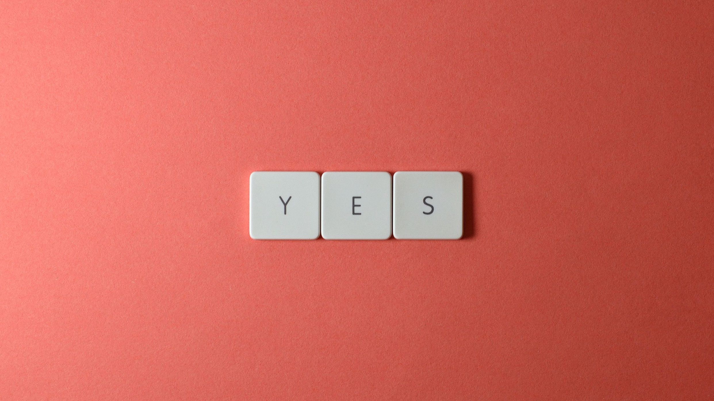 Small wooden letter tiles spelling "YES" on a neutral background, representing overcommitting and people-pleasing in marriage.
