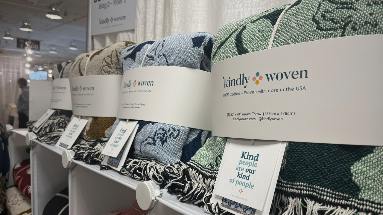 Detail of Kindly Woven blankets packaged and displayed on shelf with trade show in background.