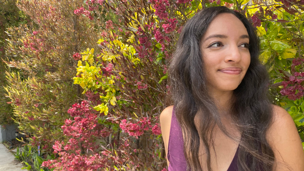 Woman in a purple dress standing outdoors in front of green foliage, looking off into the distance in a calm reflective moment