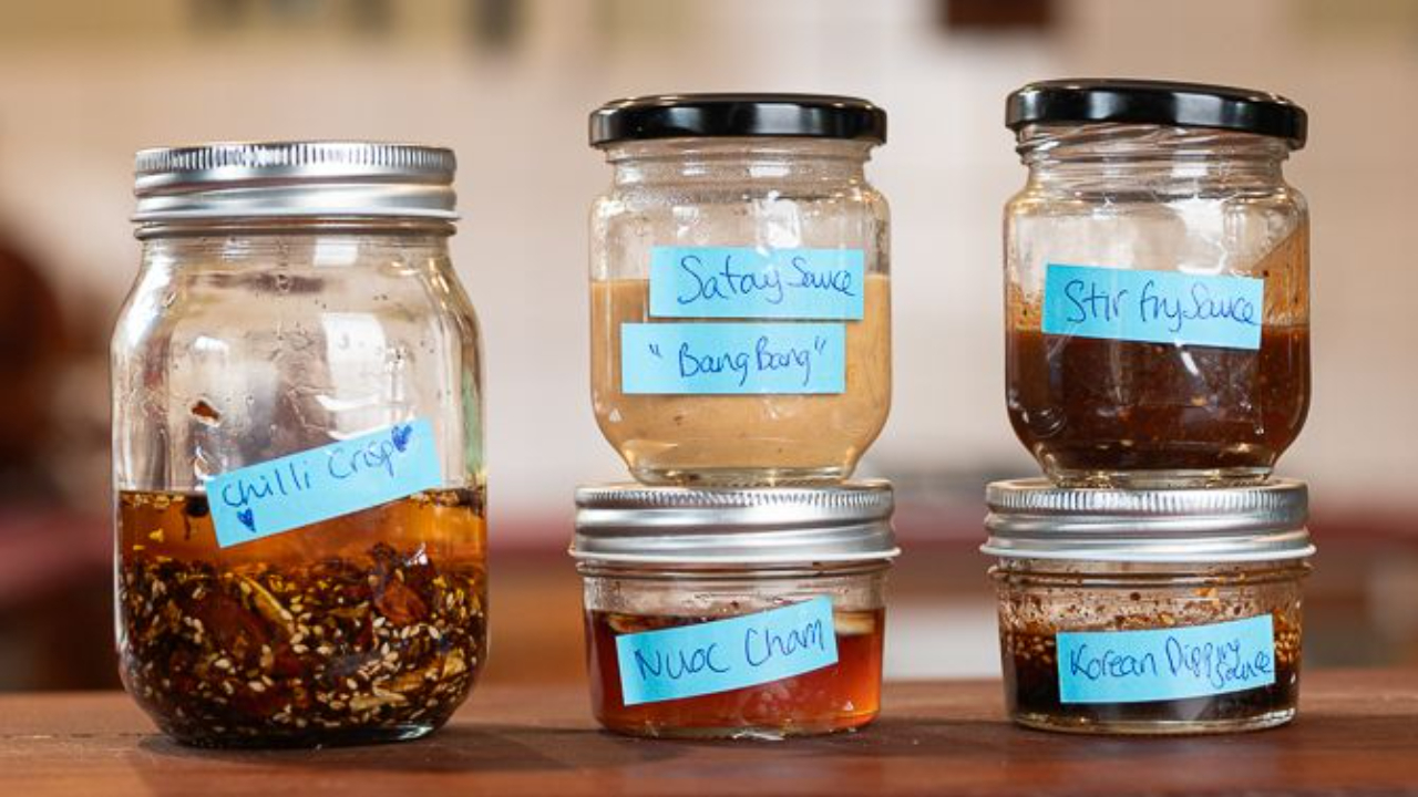 Five homemade Asian sauces in small jars and bowls on a kitchen bench, including chilli crunch, nuoc cham, satay sauce, Korean dipping sauce and stir fry sauce
