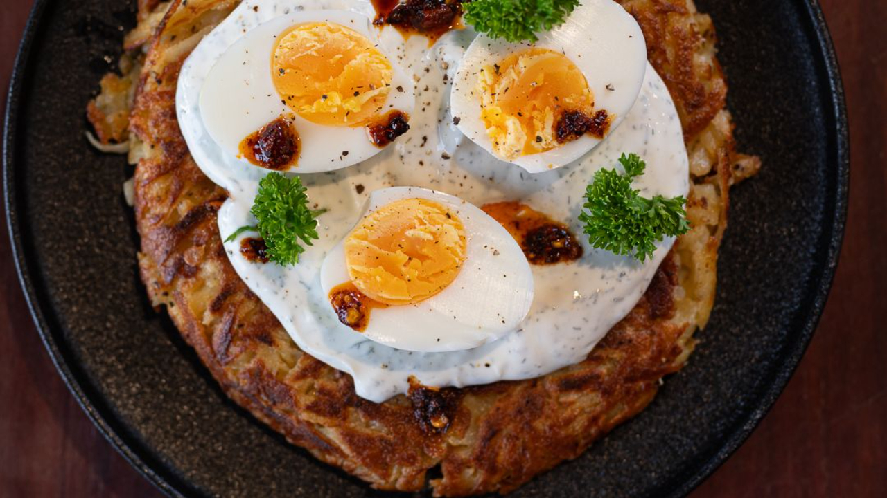 Golden crispy parsnip rosti on a plate topped with dill yoghurt, a soft boiled egg sliced open, and a drizzle of chilli crunch oil