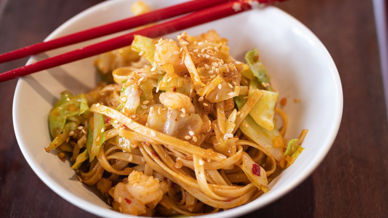A white bowl of spicy prawn noodles with cabbage and sesame seeds, chopsticks resting on top, with a jar of crushed chilli and a pack of thick noodles in the background