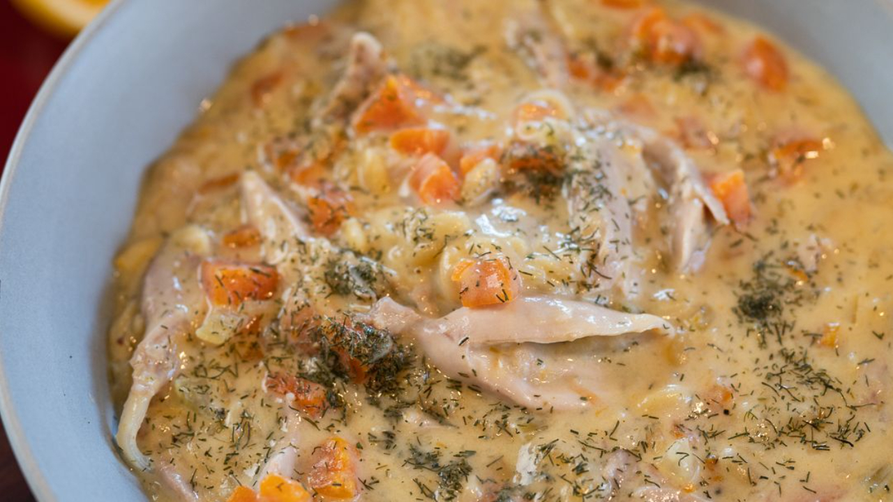 A bowl of creamy golden avgolemono Greek lemon chicken soup with shredded chicken, orzo, diced carrot and dried dill, with a squeezed lemon half and a pot of soup in the background