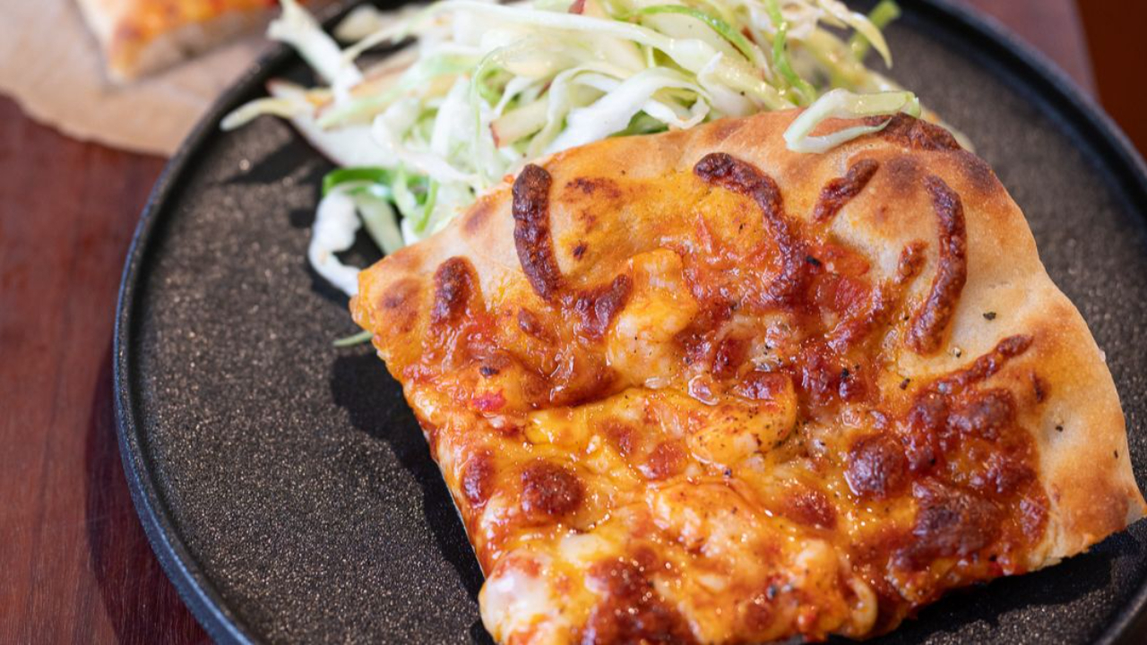 A slice of homemade spicy prawn pizza with golden bubbled cheese on a black plate, served with a fresh cabbage and apple slaw, with the full pizza on baking paper in the background
