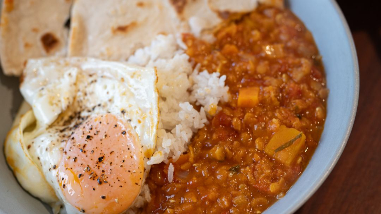 A bowl of spiced red lentil dahl with butternut squash, white rice, and a fried egg with a runny yolk, served alongside homemade flatbreads with golden brown spots