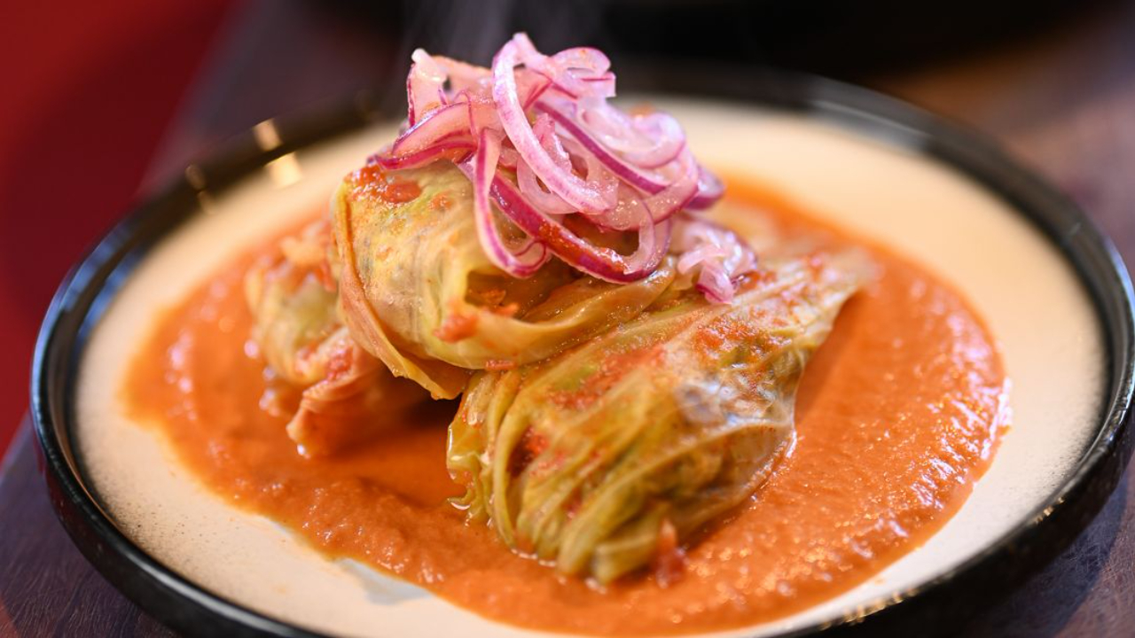  Steaming chicken cabbage rolls on a spiced tomato cream sauce in a dark rimmed bowl, topped with bright pink pickled red onions, with a pan of rolls in the background