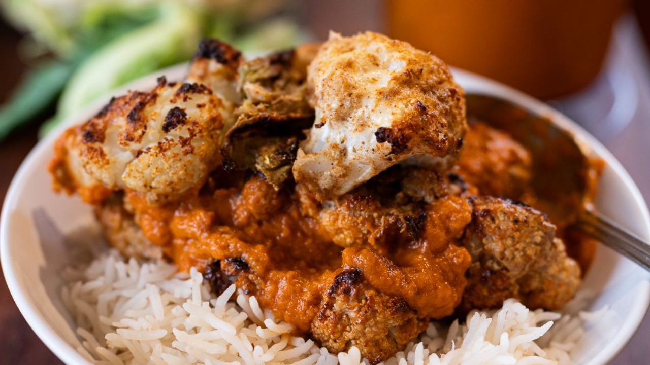 Charred cauliflower tikka masala florets on a bed of basmati rice in a white bowl, with a whole cauliflower and jar of tomato masala sauce blurred in the background
