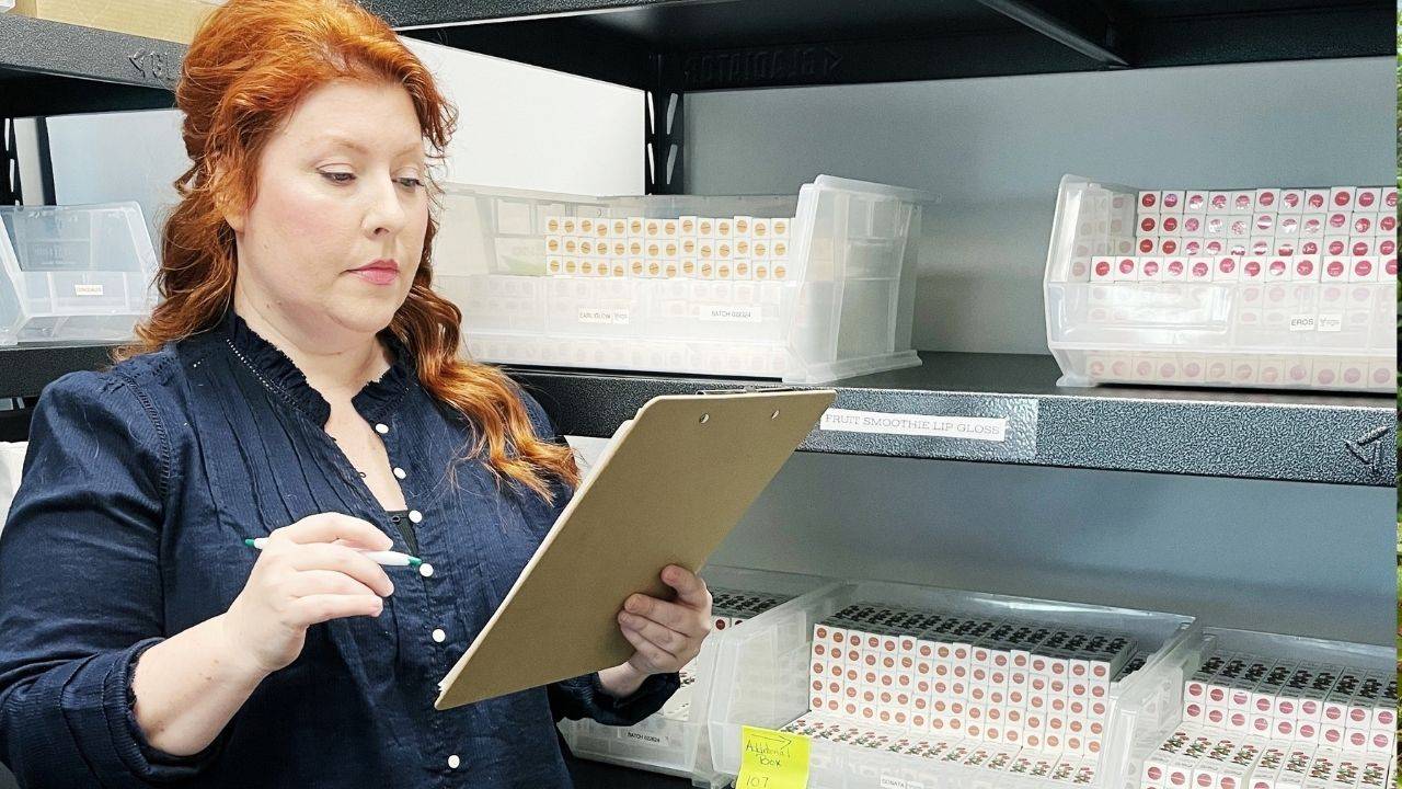 white redheaded woman stands in front of shelves full of lip gloss boxes with a clipboard in hand