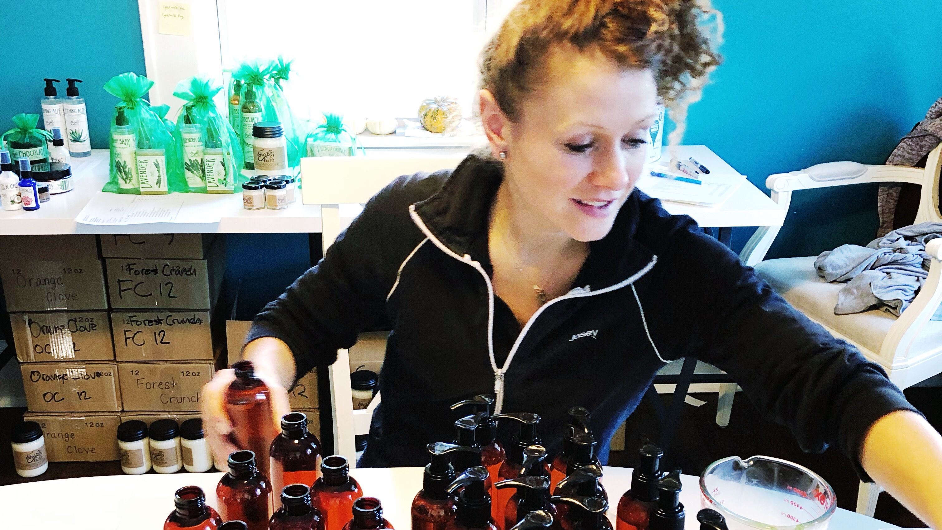 white woman smiling while bottling a body wash in a dining room with tables behind her filled with orders