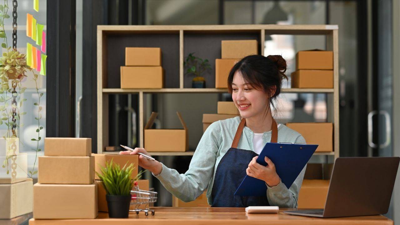 young asian woman at desk with computer, clipboard and boxes of orders she's shipping