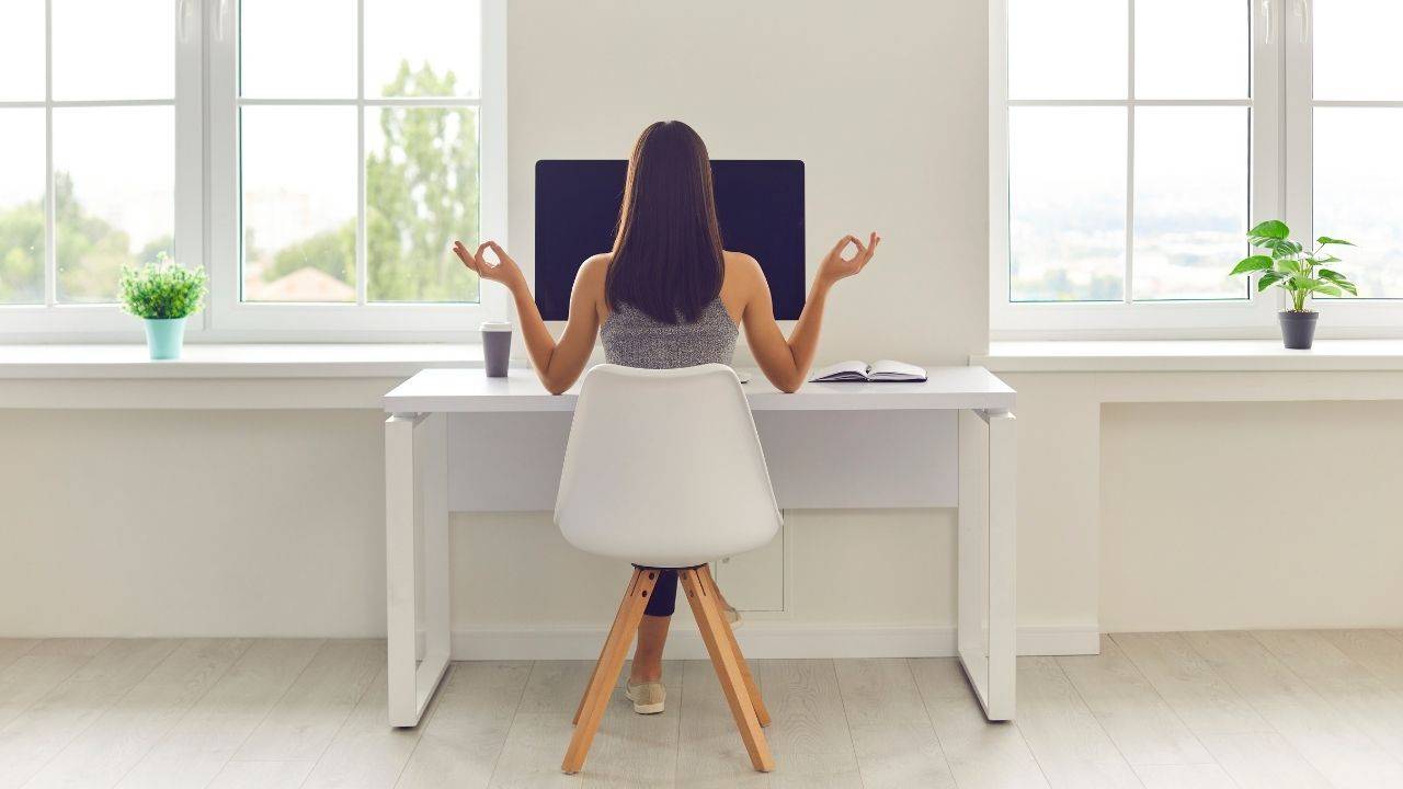 brunette woman sitting at a computer with meditating hands.  we see her from behind,  she's stitting in a white chair in front of a white desk and a white wall - a lot of white!