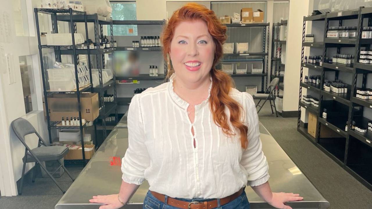 white woman with red hair in jeans and white top leaning on a table in a large room filled with shelves of beauty products