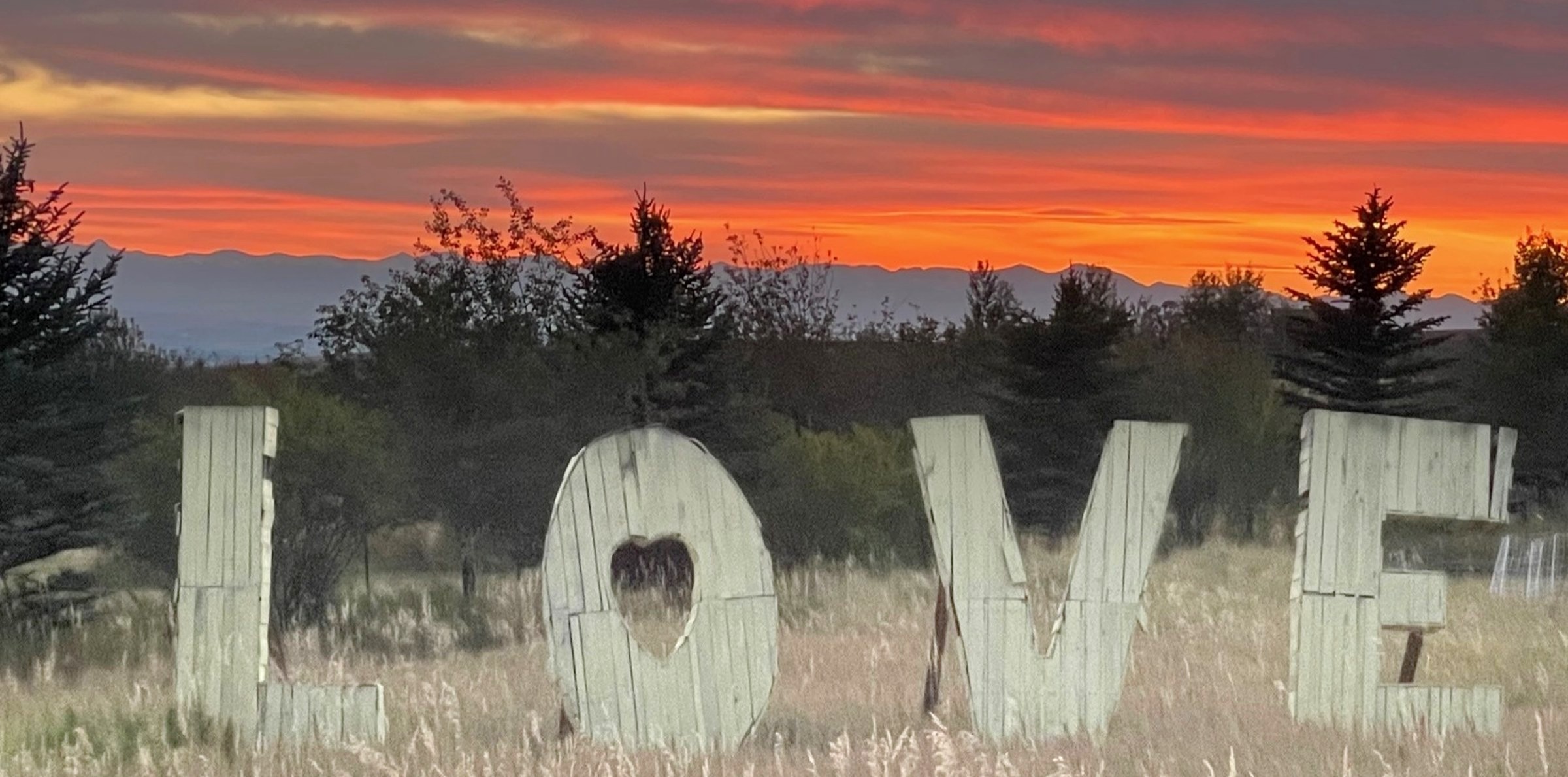 Large wooden letters spelling LOVE in a grassy field at sunset, with mountains on the horizon near Bozeman, Montana