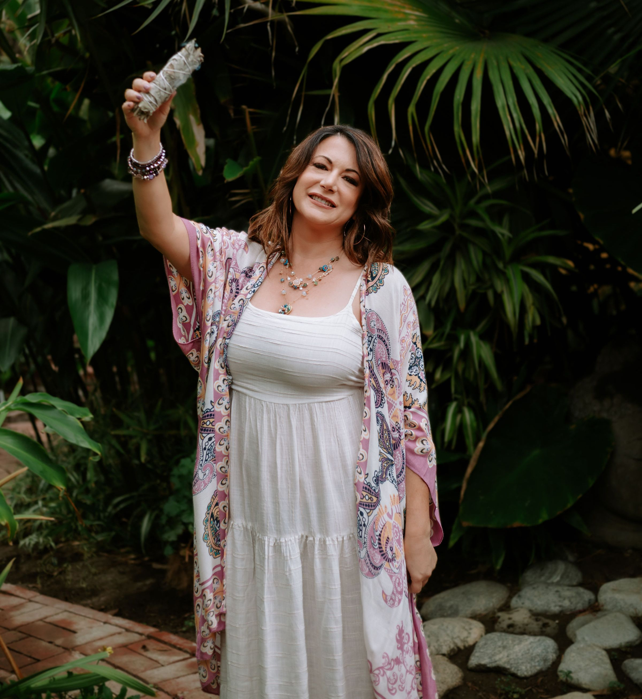 Ashley Levin in front of Palm Tree Holding a Burning Bundle of Sage and Smiling at the Camera