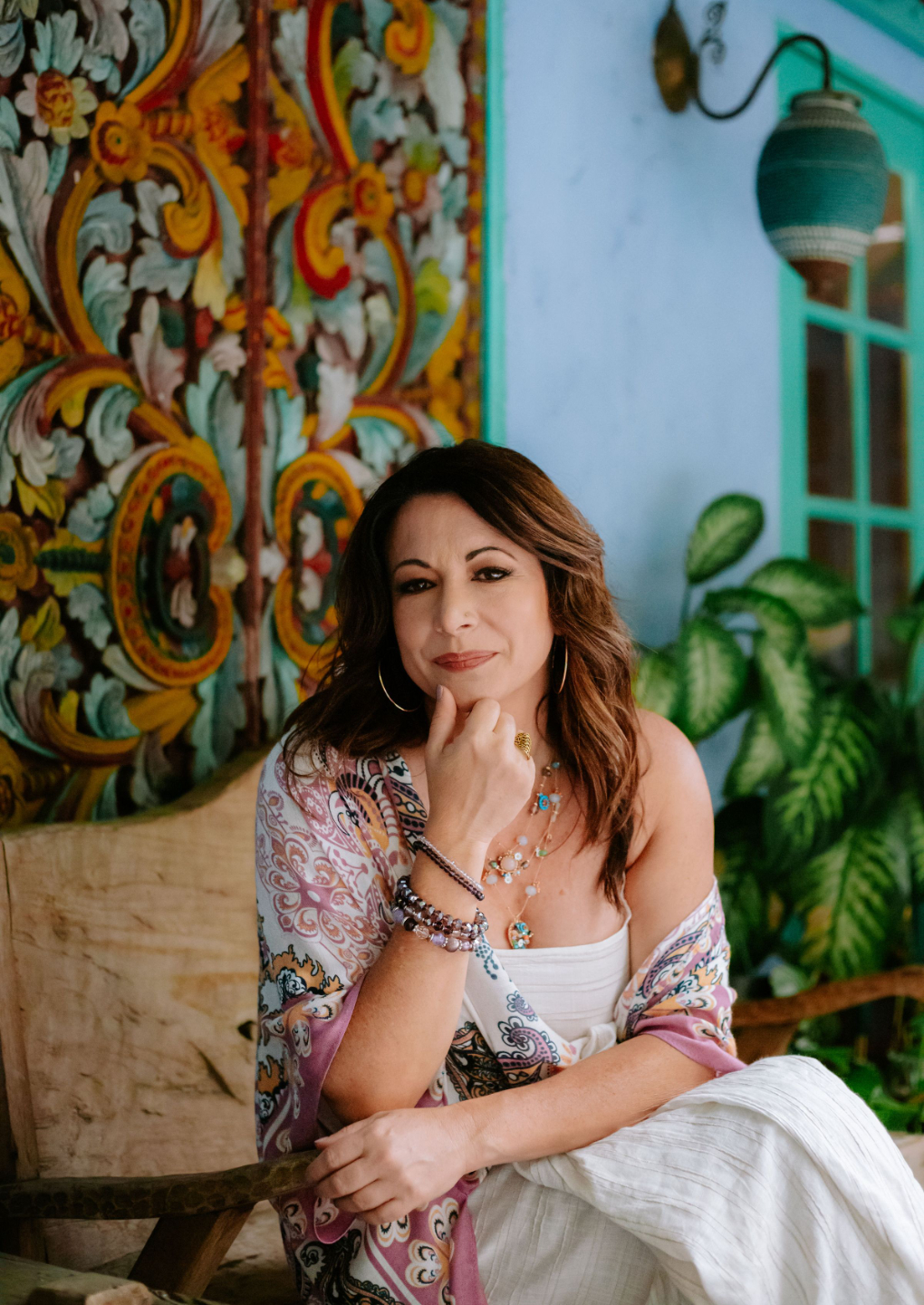 Ashley Levin Sitting on Wood Bench in front of a Colorful Turquoise Wall, Carved Wood, Wearing a White Dress and Looking at the Camera with Her Chin Resting on her Hand