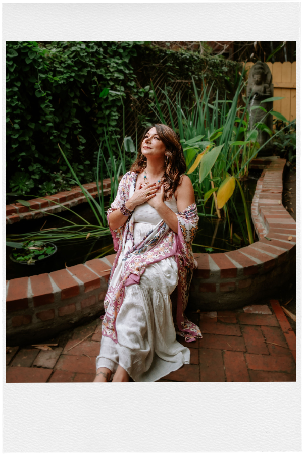 Ashley Levin Sitting on Brick, Wearing a White Dress with Her Hands on Her Heart and Looking up at the Sky