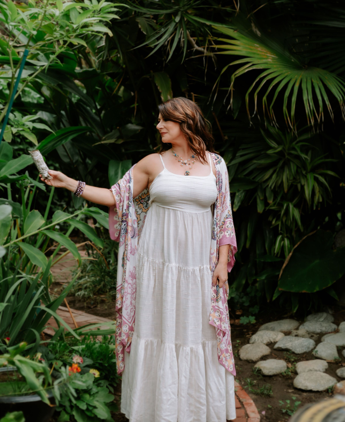 Ashley Levin Sitting on Wood Bench in front of a Colorful Turquoise Wall, Carved Wood, Wearing a White Dress and Looking at the Camera with Her Chin Resting on her Hand
