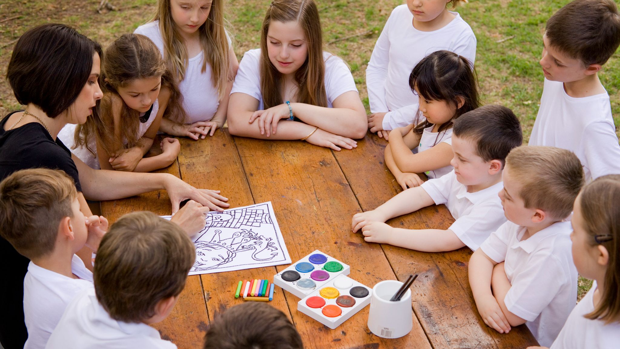 Children gathered around a rustic wooden table participating in a step-by-step online art lesson with watercolour paints and drawing materials outdoors.
