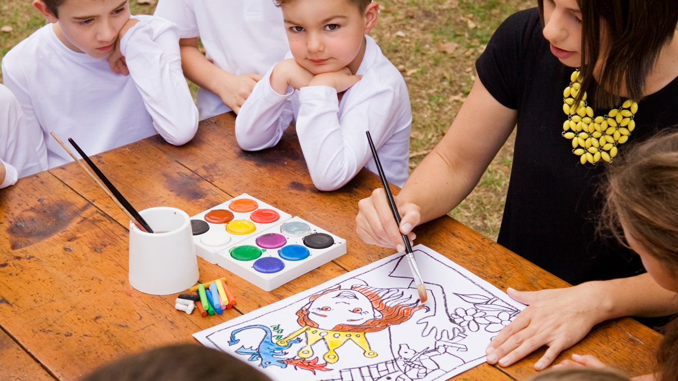 Children watching an artist demonstrate a colourful step-by-step drawing lesson with watercolour paints on a rustic wooden table outdoors.