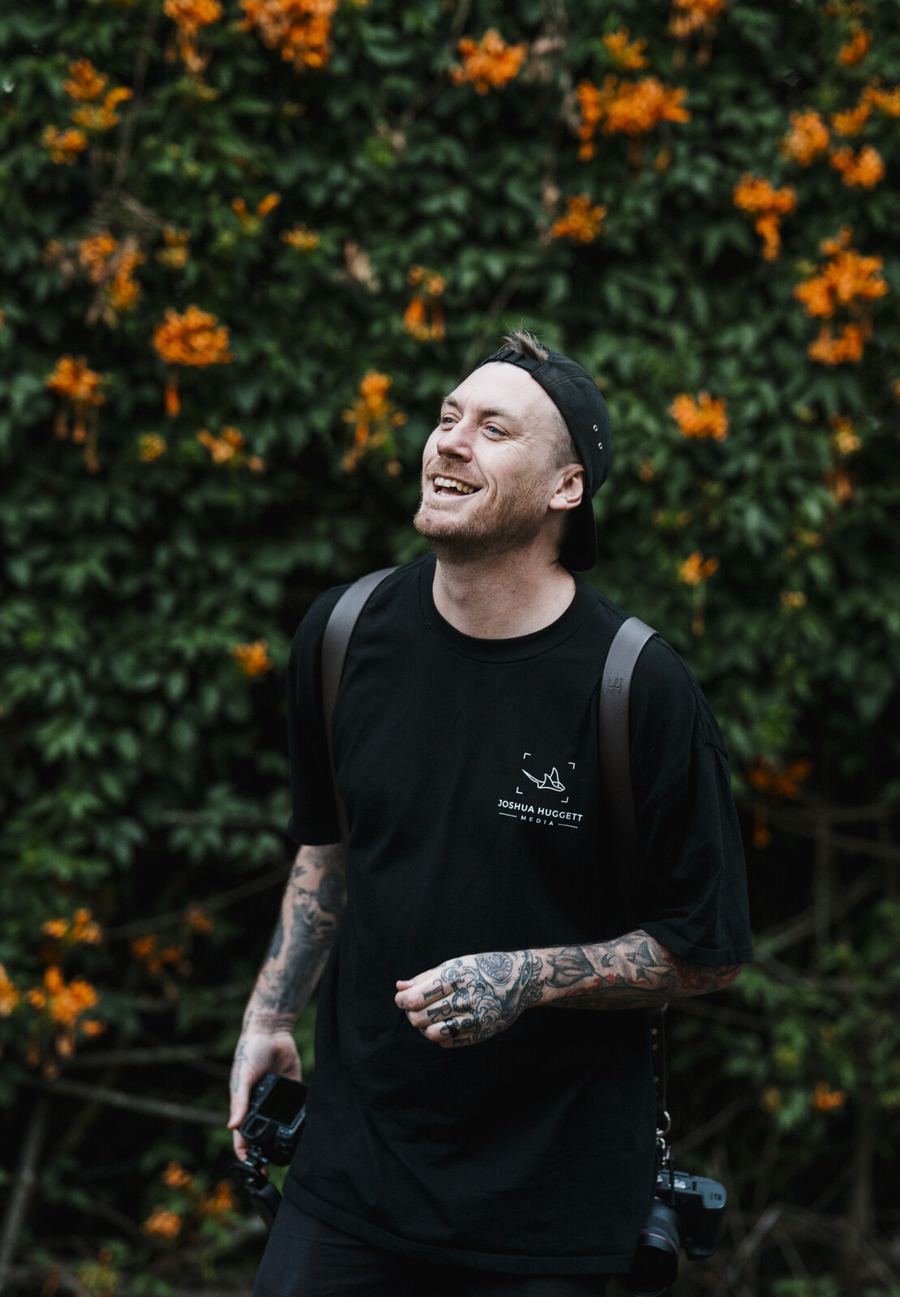 Josh Huggett laughing, camera gear on, in front of orange flowers at a wedding