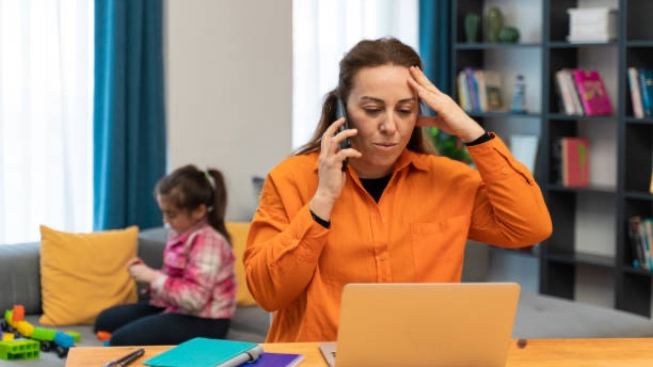 Stressed mom sits at working desk behind computer while talking on the phone.