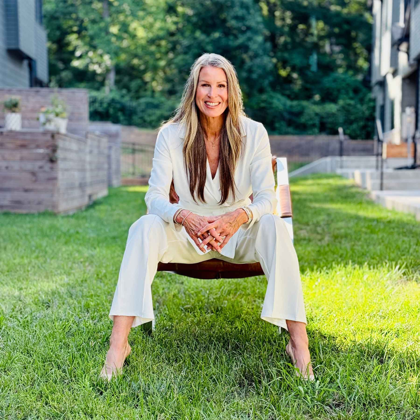 Long-haired woman in a pants suit sitting with hands folded, leaning forward in her outdoor chair.