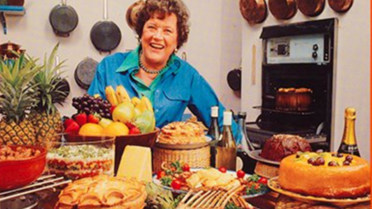Julia Child surrounded by food with an oven and pots and pans on the walls behind her.