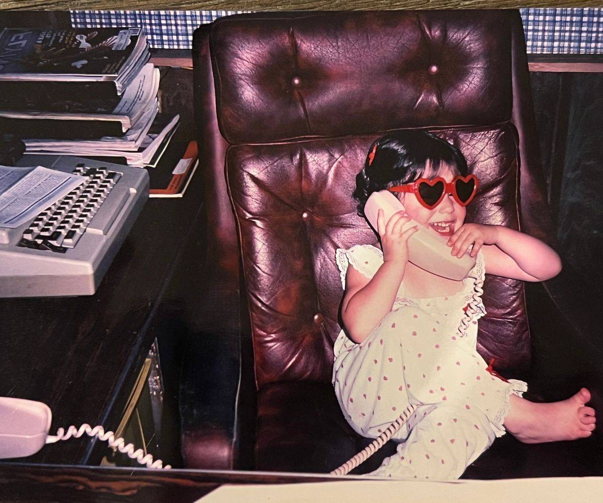Toddler Tiffany in heart-shaped sunglasses, talking on the phone from a big leather office chair.