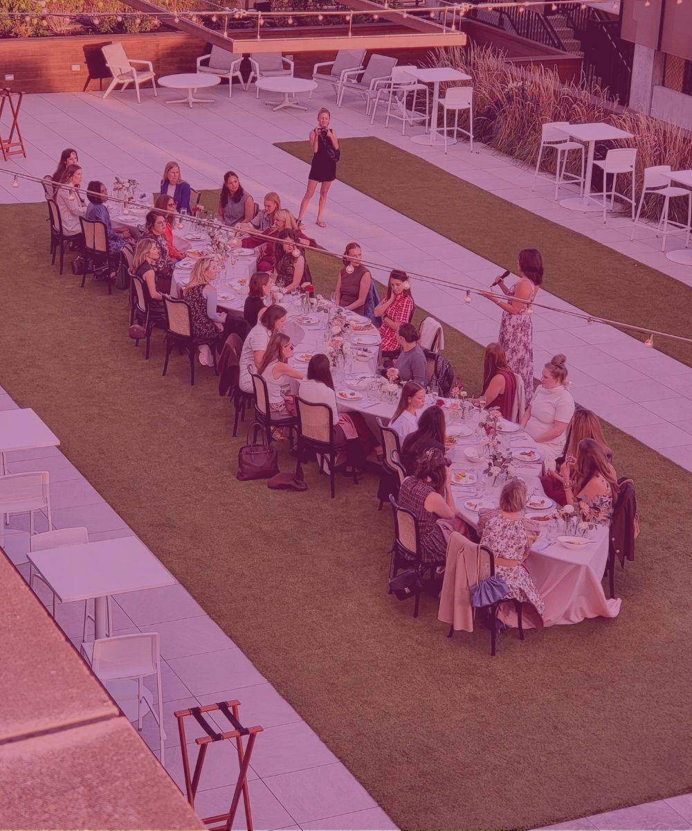 Seat at the Table 2026: women seated along one long dinner table on a rooftop lawn as a host speaks from the end of the table.