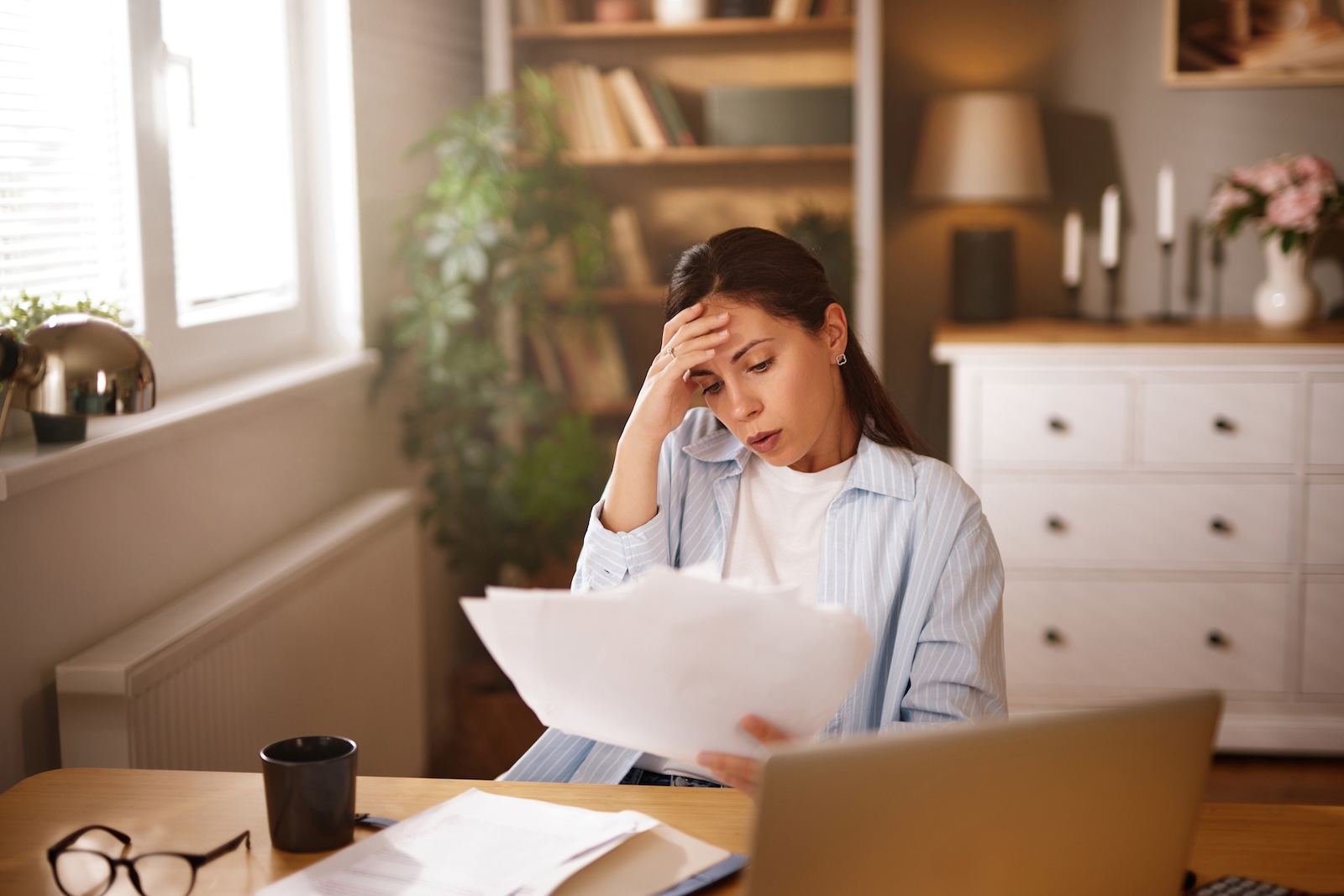Worried woman at a home desk reading official paperwork beside her laptop