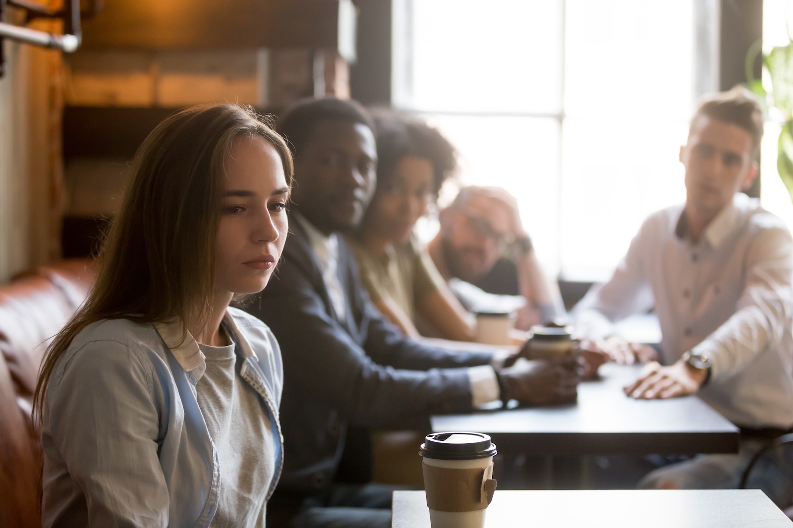 Woman sitting alone with a coffee cup in a café while a group at the table behind her looks on