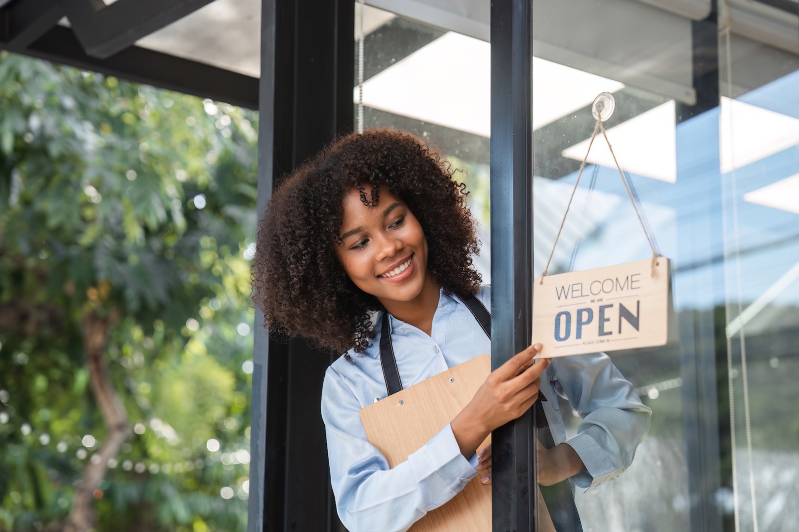 Smiling woman with a clipboard turns a "Welcome, we are open" sign on a café door