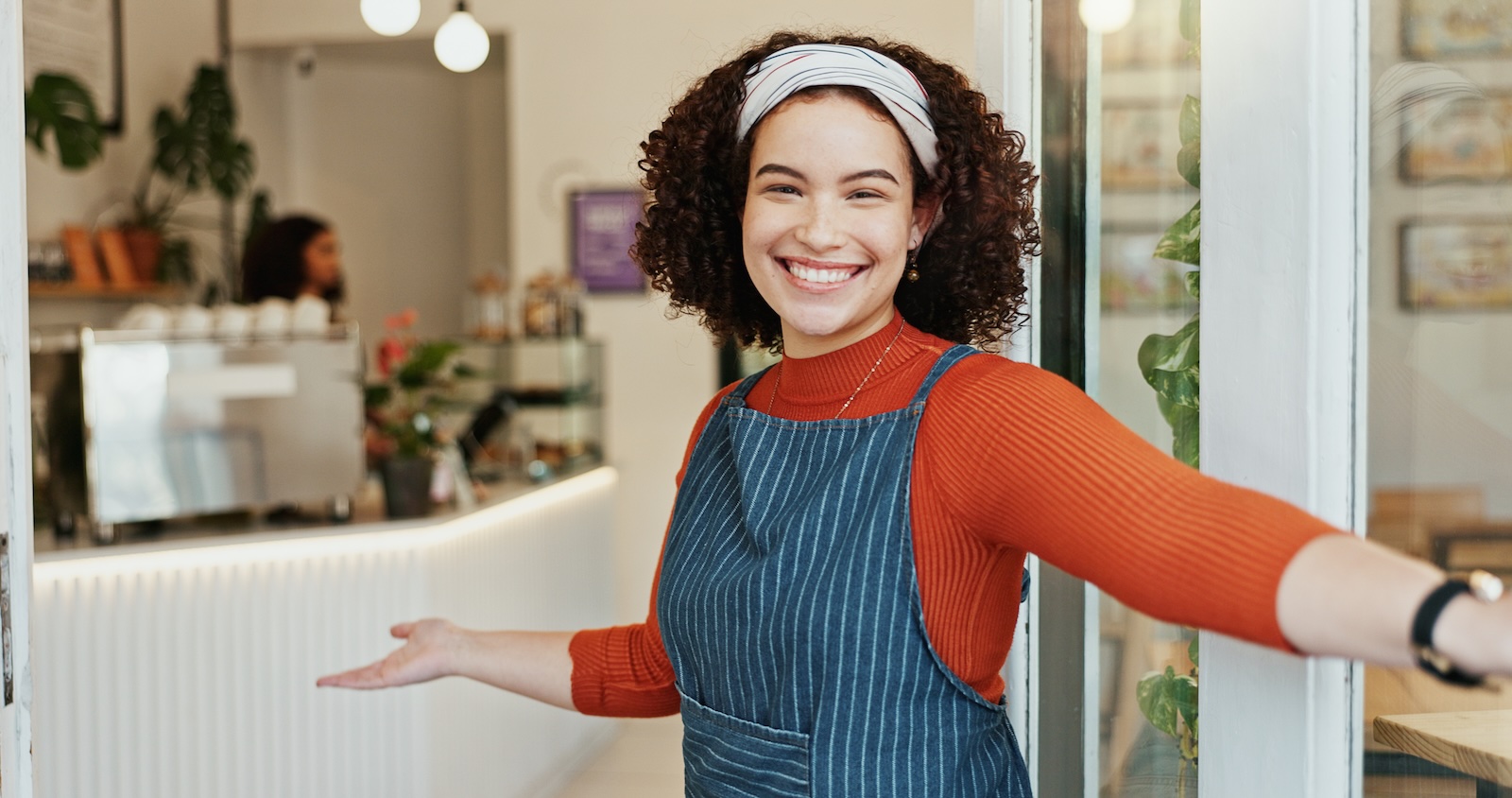 Smiling café worker in a striped apron opens the door with open arms in a warm welcome