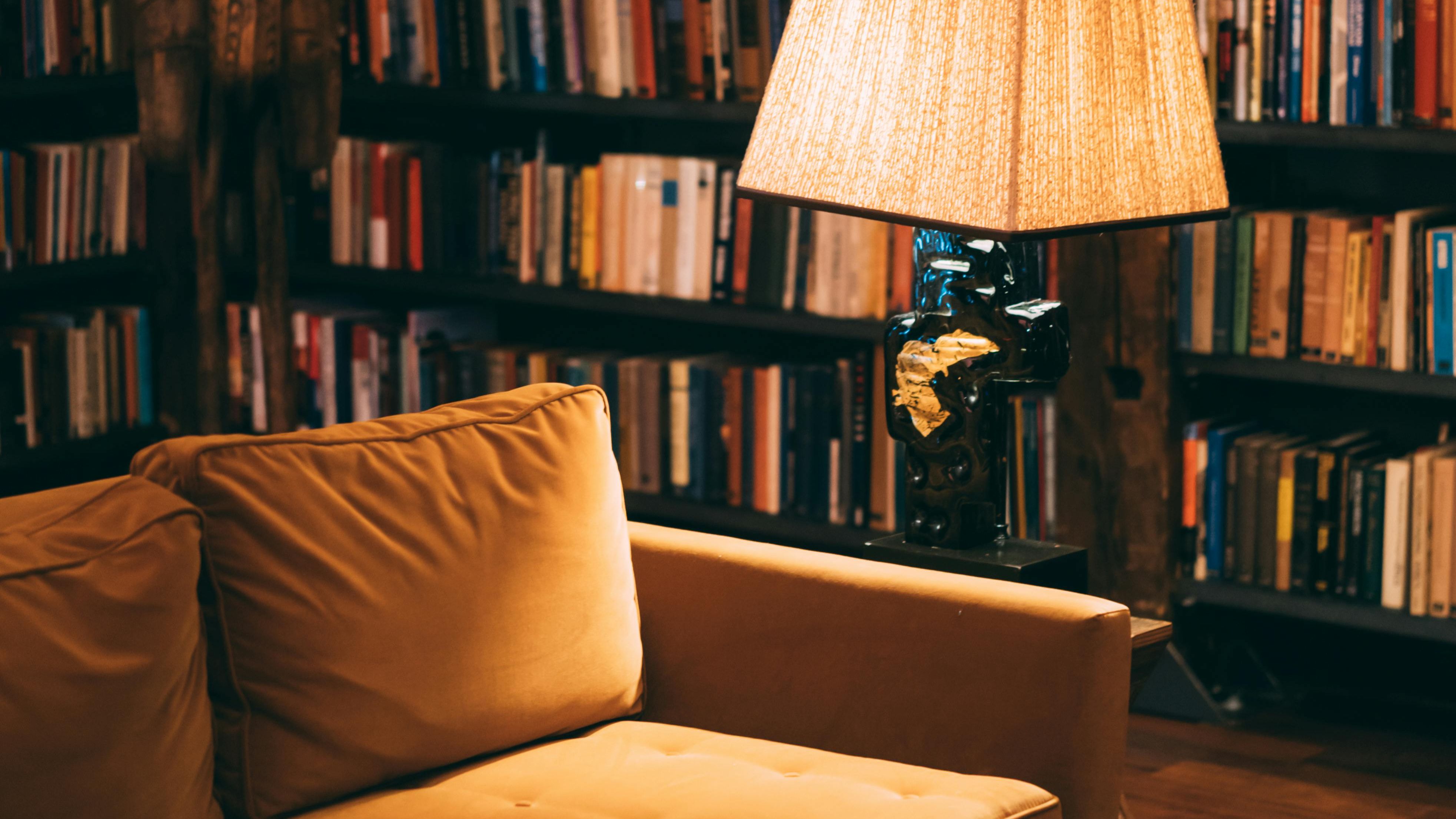 Warm, softly lit library interior with floor-to-ceiling bookshelves, a mustard sofa, and a reading lamp, creating a reflective and thoughtful atmosphere.