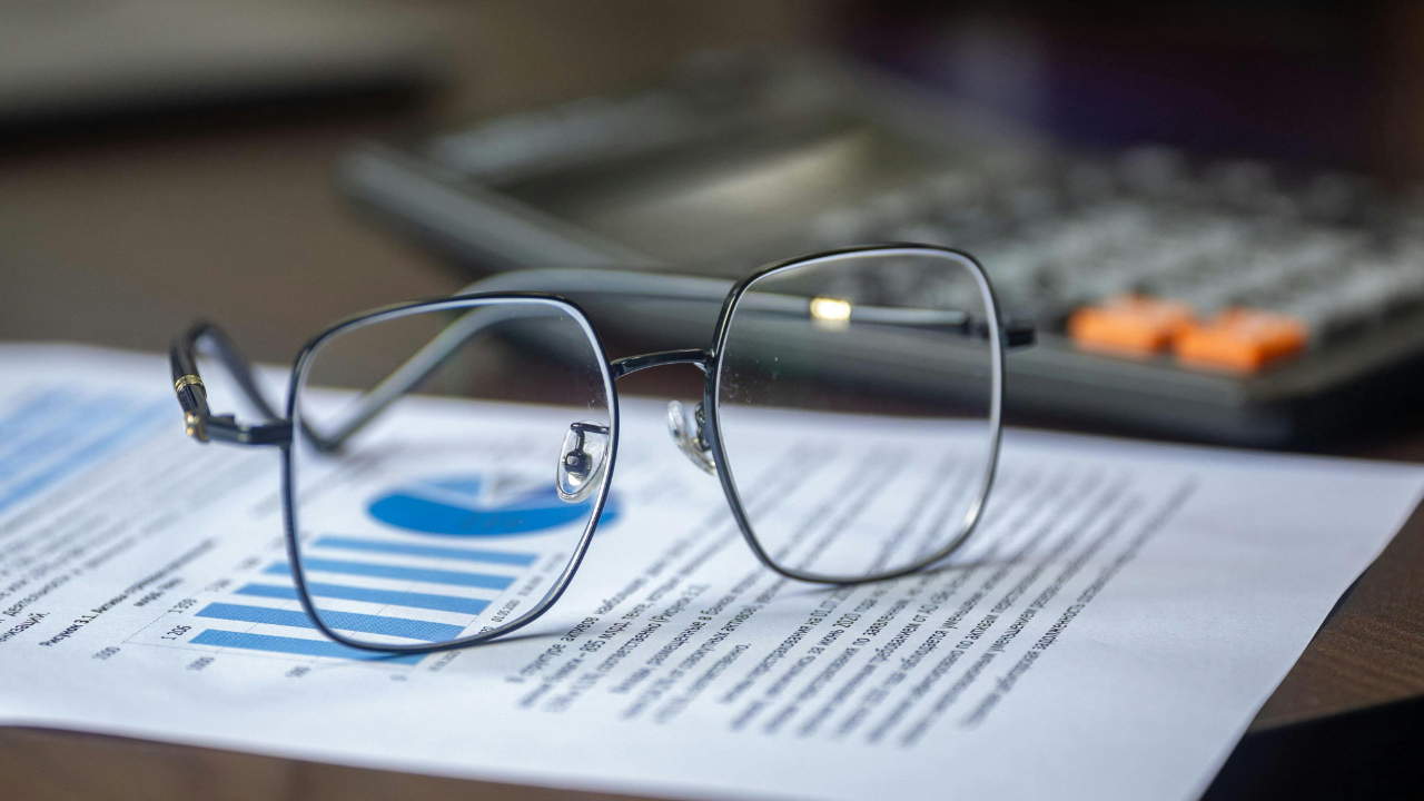 Close-up of eyeglasses resting on financial documents with charts and graphs, with a calculator blurred in the background, suggesting compensation analysis and career planning.