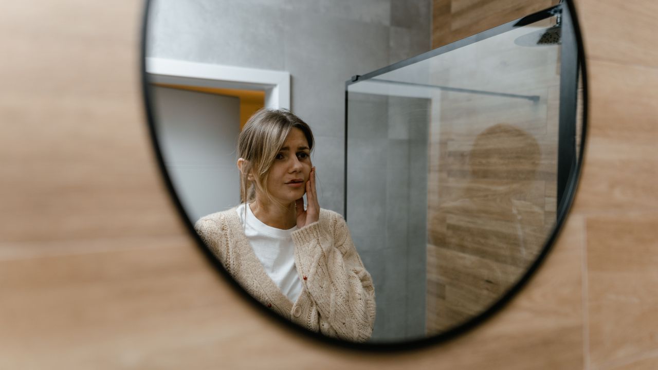 Woman looking at her reflection in a round mirror with a worried expression, noticing her inner critic