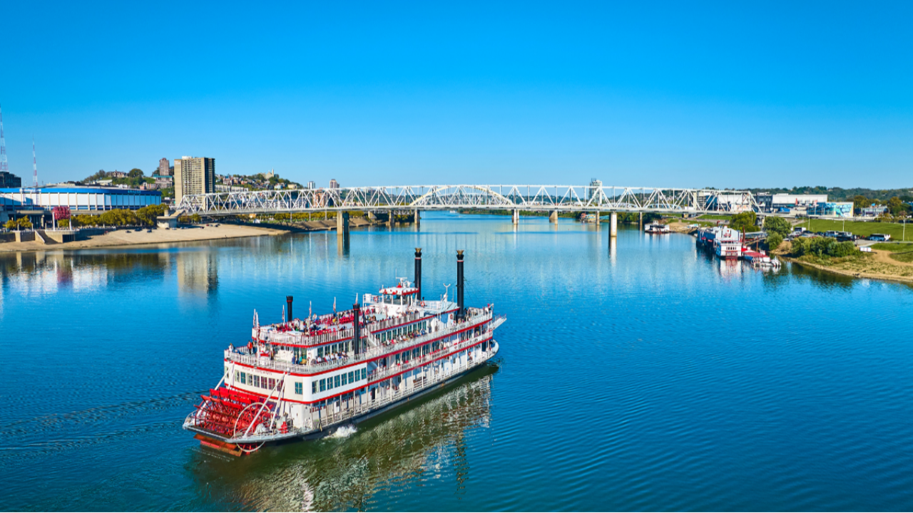 A riverboat cruises along the Ohio River with a Cincinnati-area skyline, bridges, and waterfront visible in the background.