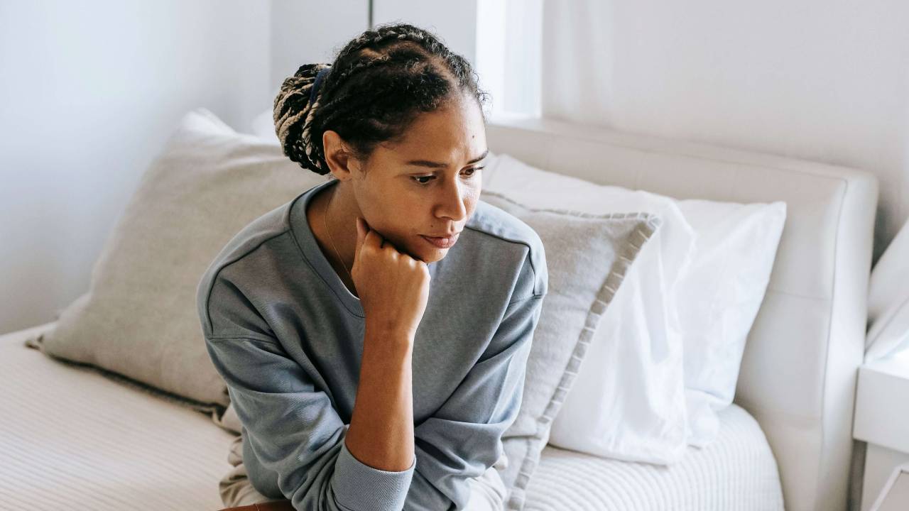 Woman with braided hair in a grey sweatshirt sitting on a white bed resting her chin on her fist with a thoughtful distant expression | international trauma therapist | therapeutic coaching | executive coaching | Seattle | New York City | Chicago