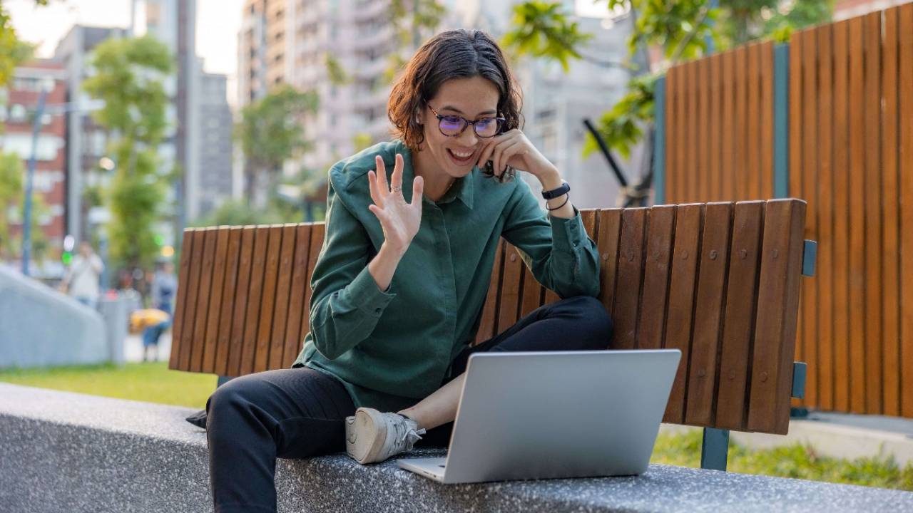Woman with glasses and short hair smiling and waving at a laptop screen while sitting on an outdoor bench in a city | therapeutic coaching online | international trauma therapist | therapy vs coaching | New York City | Chicago | Seattle